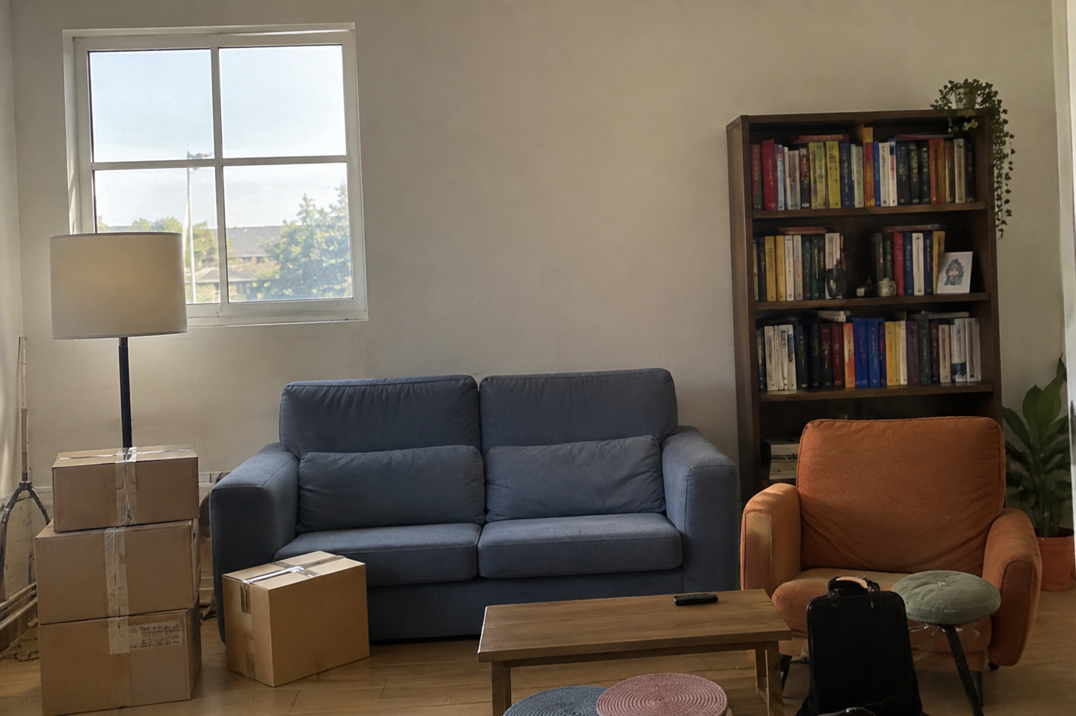 A cluttered living room with a sofa, armchair, bookshelf, lamp and moving boxes